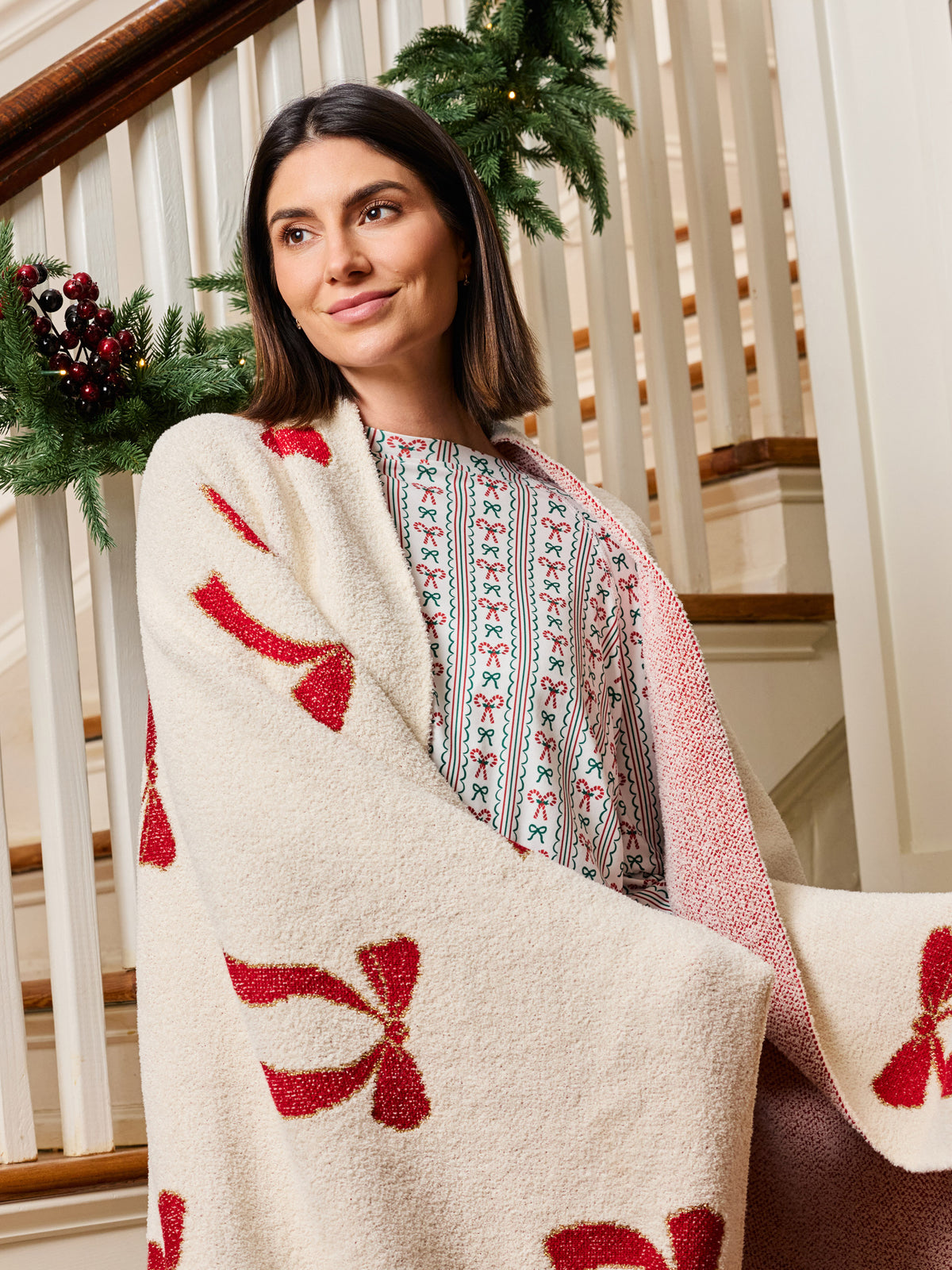 A woman wrapped in a cream knit blanket with red bow patterns, standing on a decorated indoor staircase.