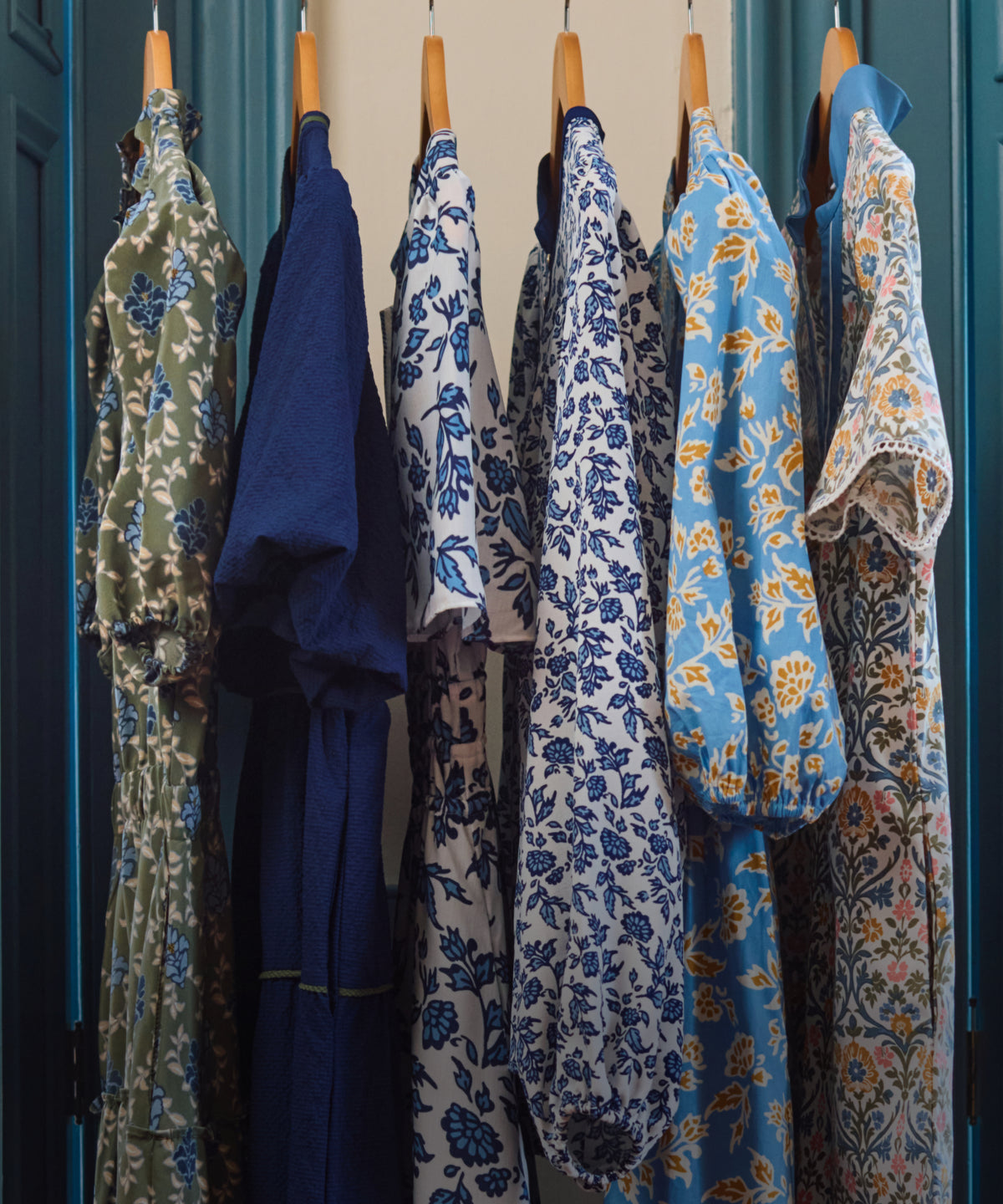 A row of long-sleeve floral and patterned dresses hanging on wooden hangers against a teal door and cream wall.