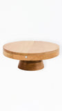 A round wooden cake stand or pedestal table with a light tan top and a darker brown base, shown against a plain white background.
