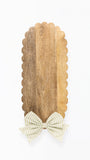A vertical wooden serving board with a scalloped edge and a striped bow at the bottom, shown on a plain white background.
