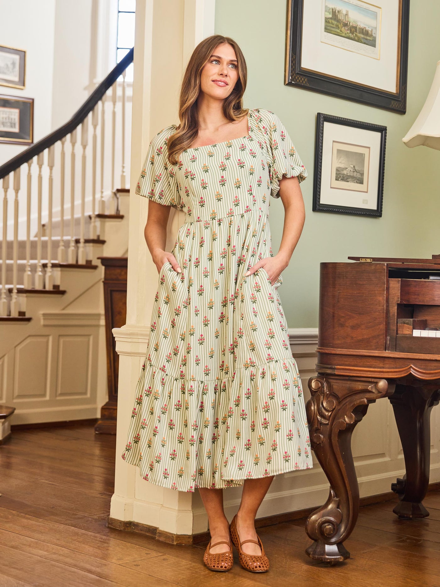 A woman standing in an interior room wearing a green floral dress with a square neckline and puff sleeves.