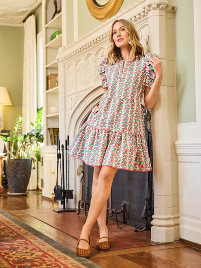A woman standing in a room with elegant decor, wearing a short-sleeved knee-length dress with a geometric pattern in shades of pink and yellow, paired with a belt, and beige strappy sandals.