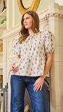A woman wearing a beige top with a floral pattern, short puff sleeves, and elastic cuffs, paired with blue jeans.