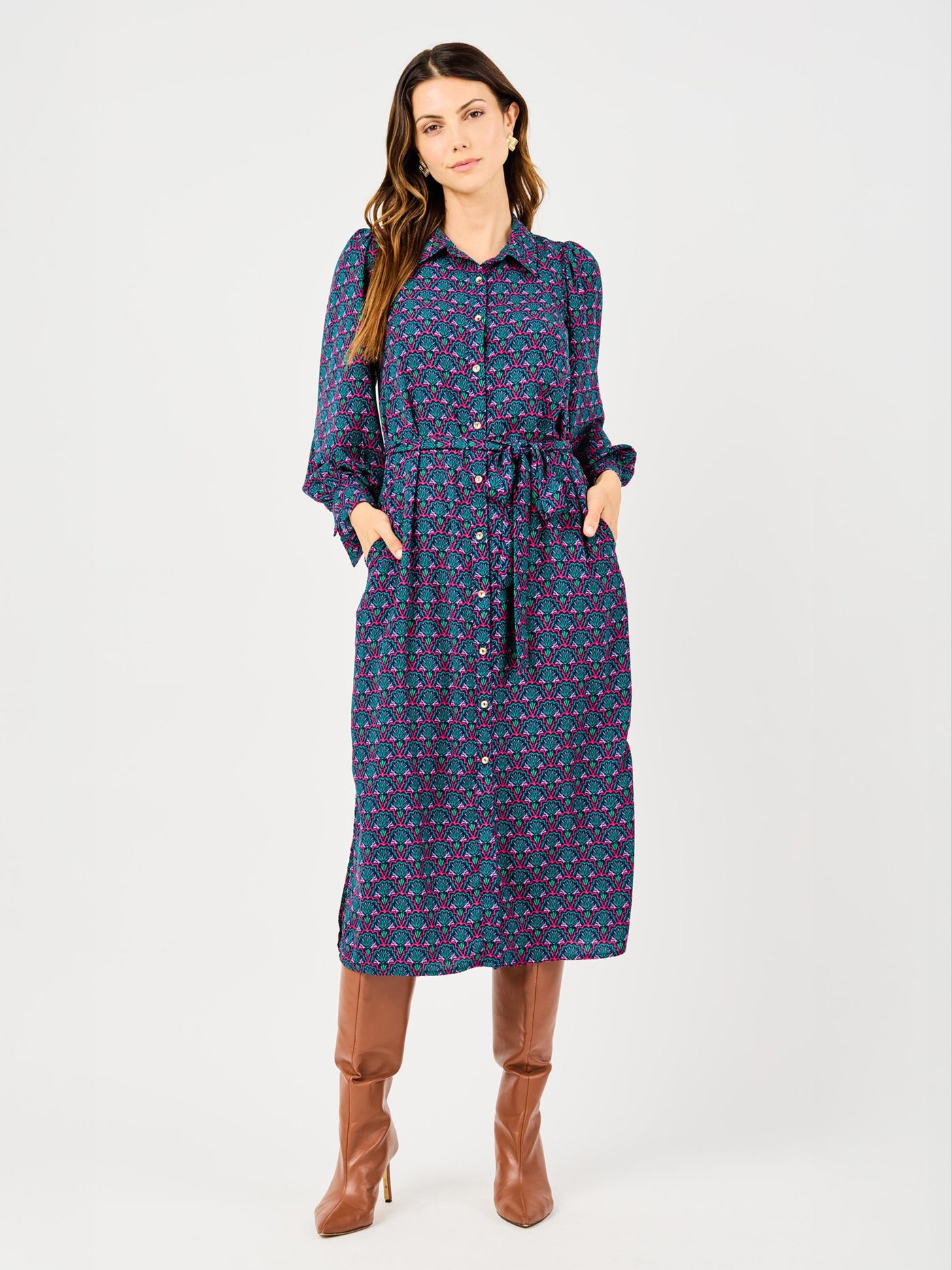 A woman standing against a white background wearing a long sleeve, midi-length dress with a geometric pattern, button details, and a removable belt. She is also wearing brown boots.