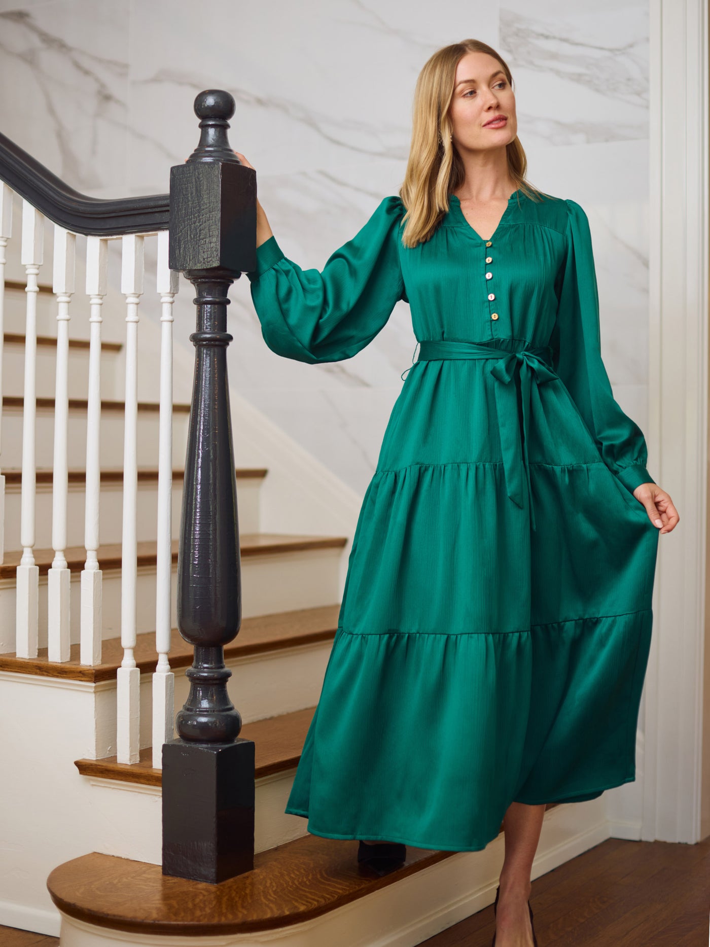 Woman in an elegant green dress with long sleeves, a tie belt and black heels standing on a staircase