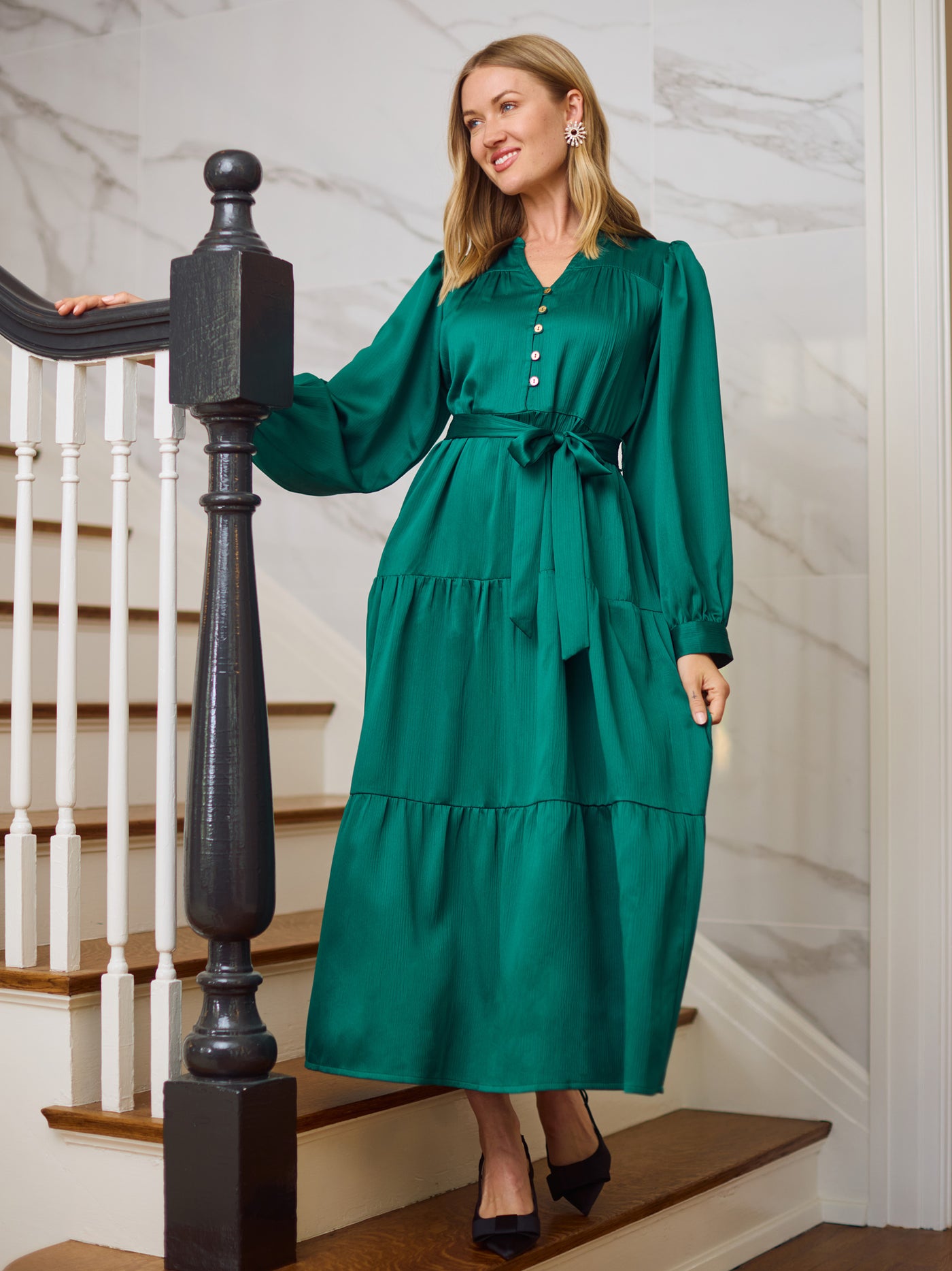 Woman in an elegant green dress with long sleeves, a tie belt and black heels standing on a staircase