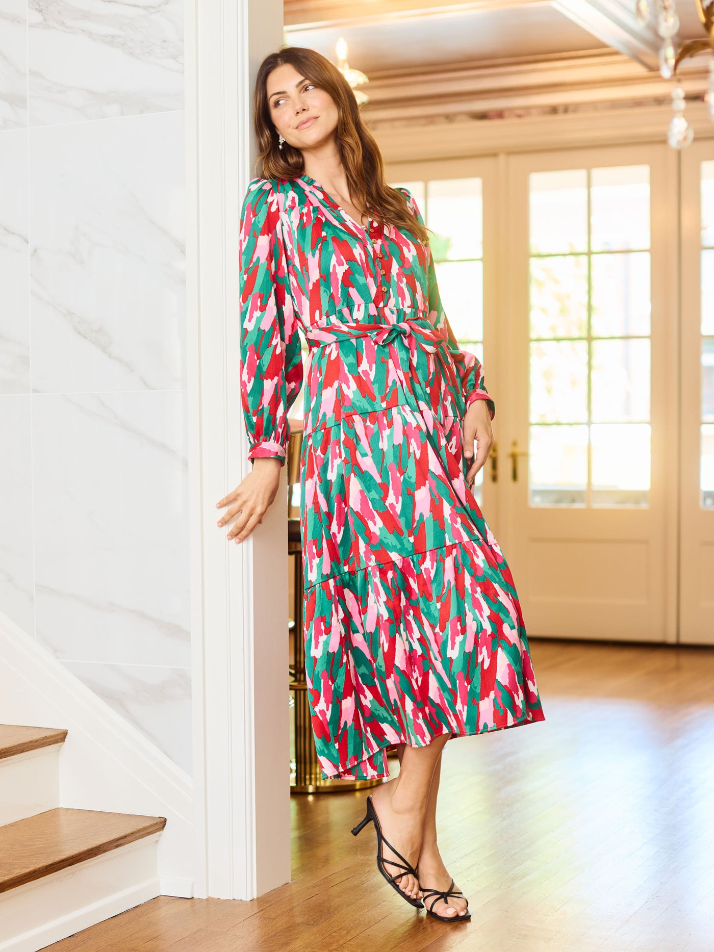 Woman wearing a colorful patterned holiday dress with pinks, greens, reds and whites leaning on a wall