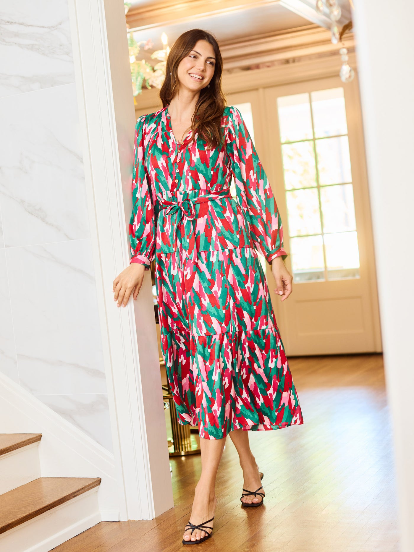 Woman wearing a colorful patterned holiday dress with pinks, greens, reds and whites leaning on a wall