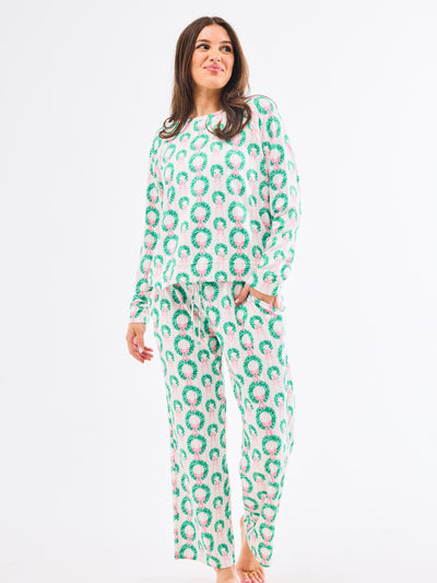 Woman wearing a green and pink patterned pajamas that feature christmas wreaths with pink bows on themon a white background