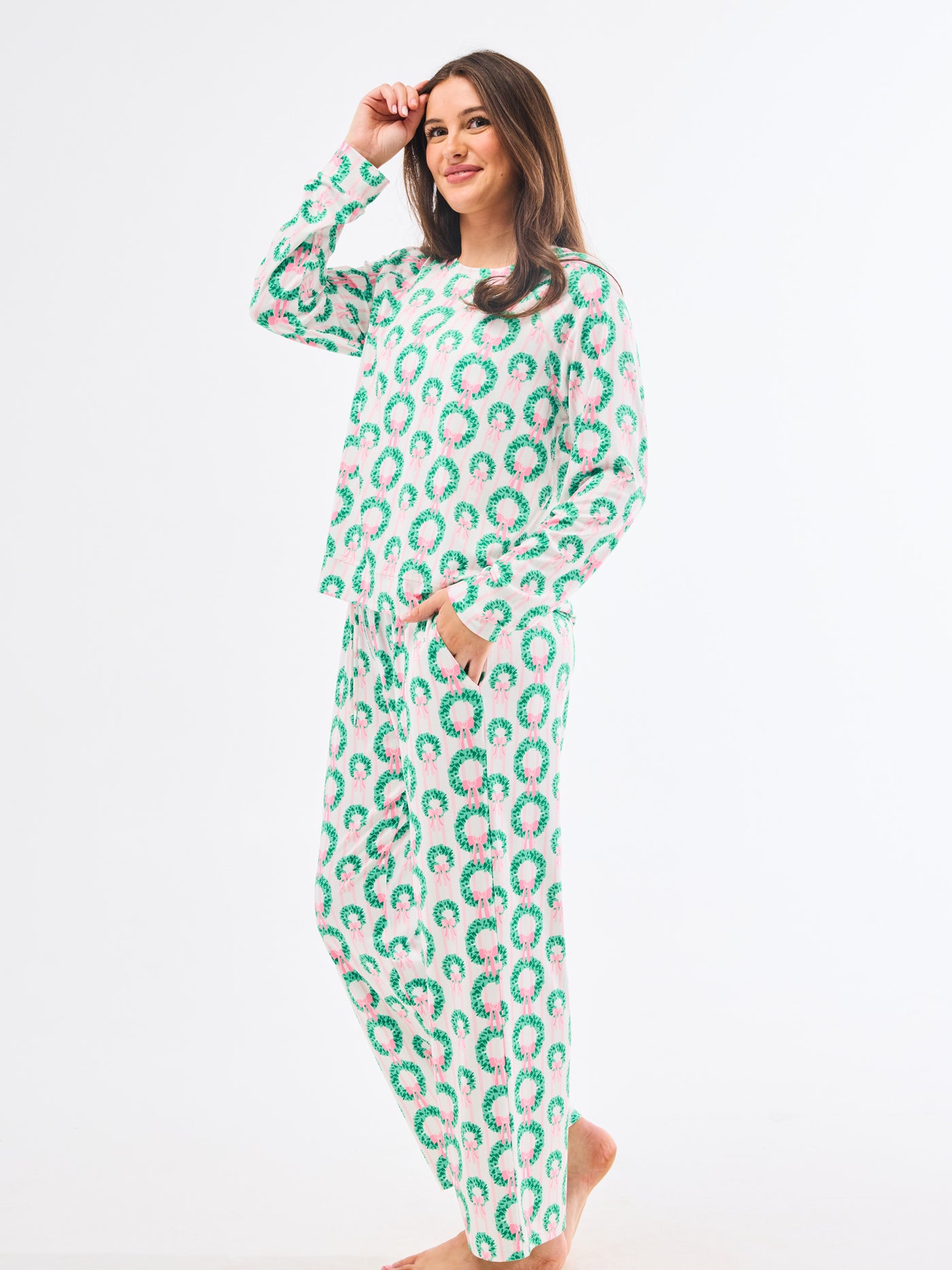 Woman wearing a green and pink patterned pajamas that feature christmas wreaths with pink bows on themon a white background