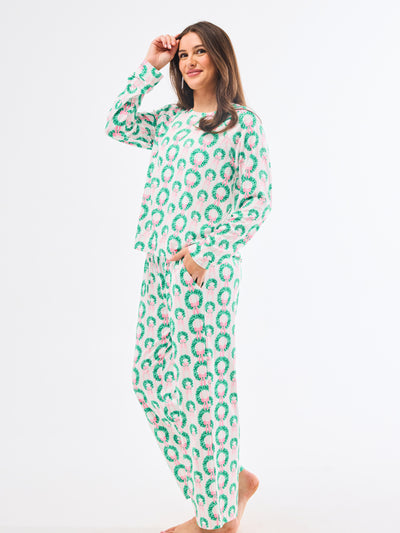 Woman wearing a green and pink patterned pajamas that feature christmas wreaths with pink bows on themon a white background