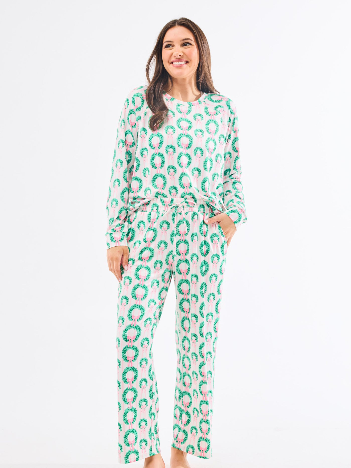 Woman wearing a green and pink patterned pajamas that feature christmas wreaths with pink bows on themon a white background