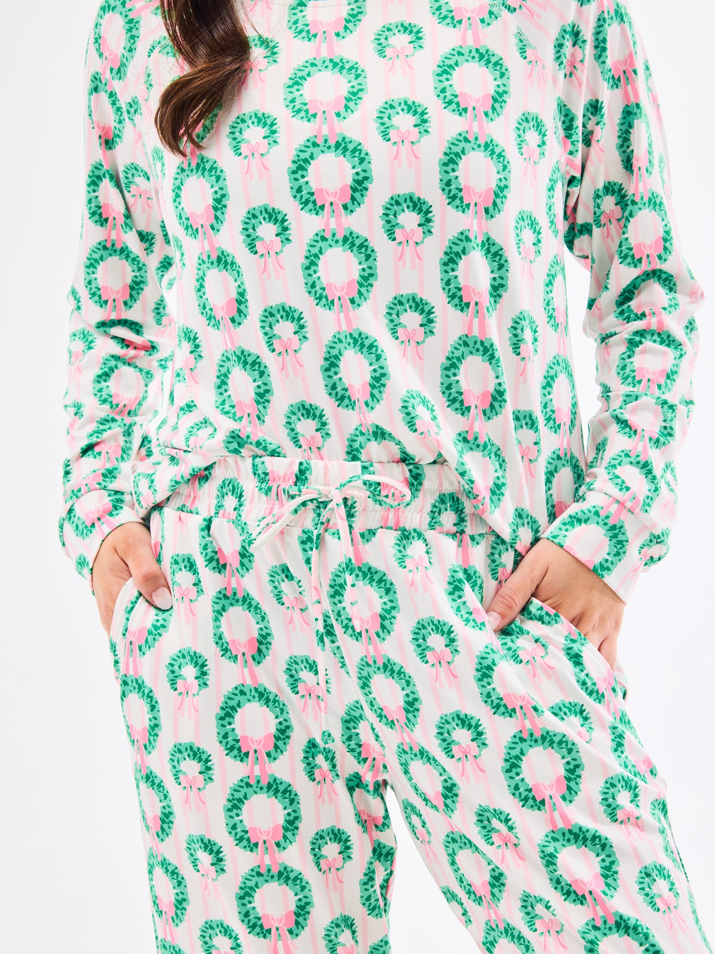 Woman wearing a green and pink patterned pajamas that feature christmas wreaths with pink bows on themon a white background