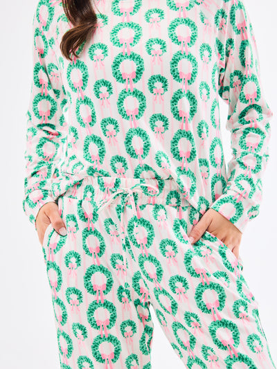 Woman wearing a green and pink patterned pajamas that feature christmas wreaths with pink bows on themon a white background