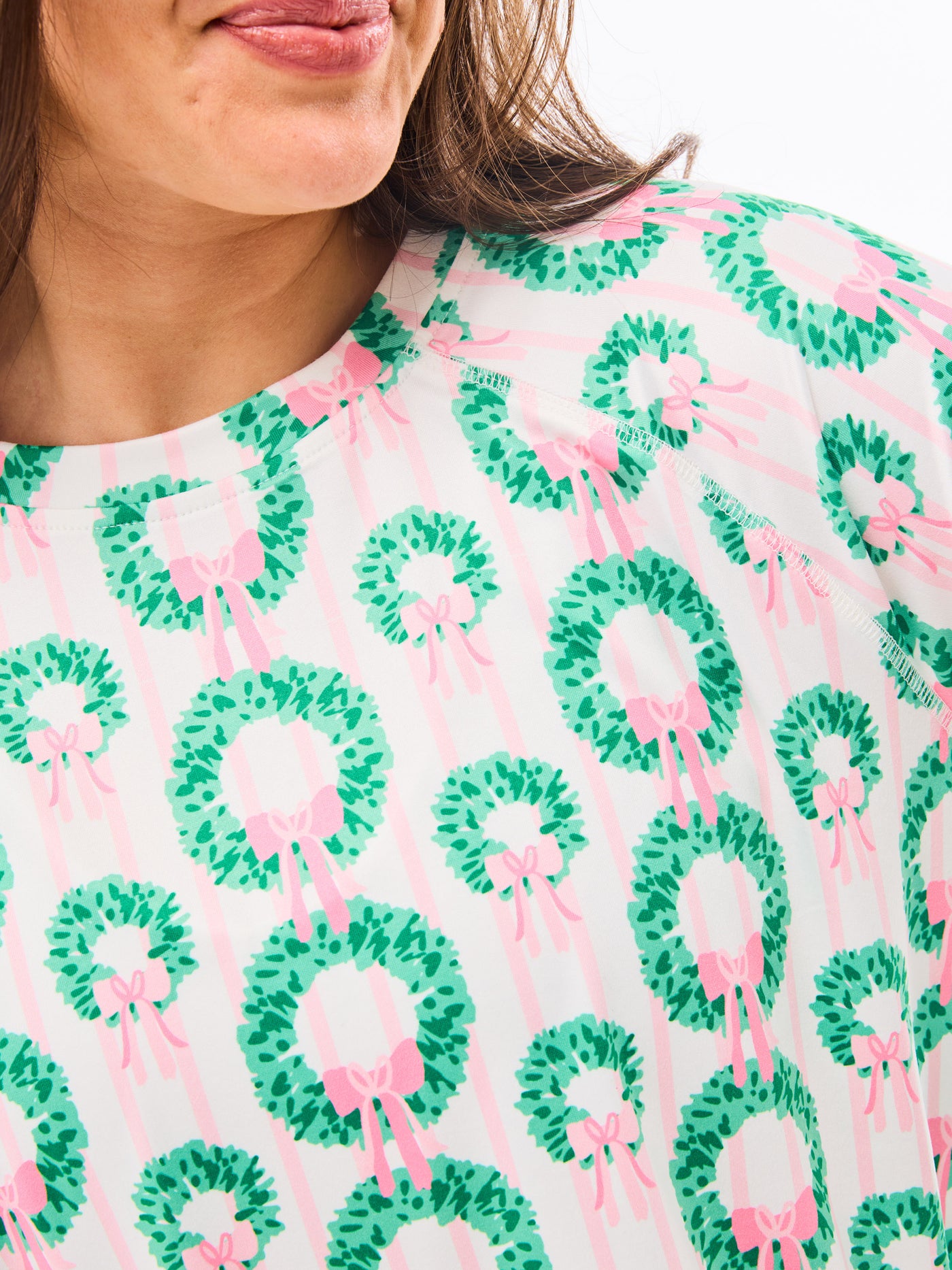 Woman wearing a green and pink patterned pajamas that feature christmas wreaths with pink bows on themon a white background