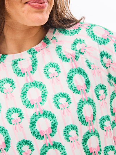 Woman wearing a green and pink patterned pajamas that feature christmas wreaths with pink bows on themon a white background