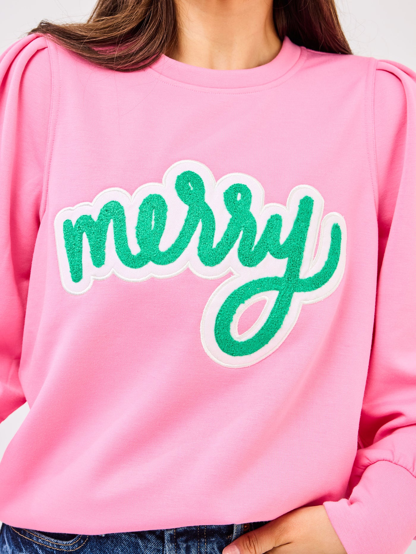 Person wearing a pink sweatshirt with 'merry' in green text on a plain background