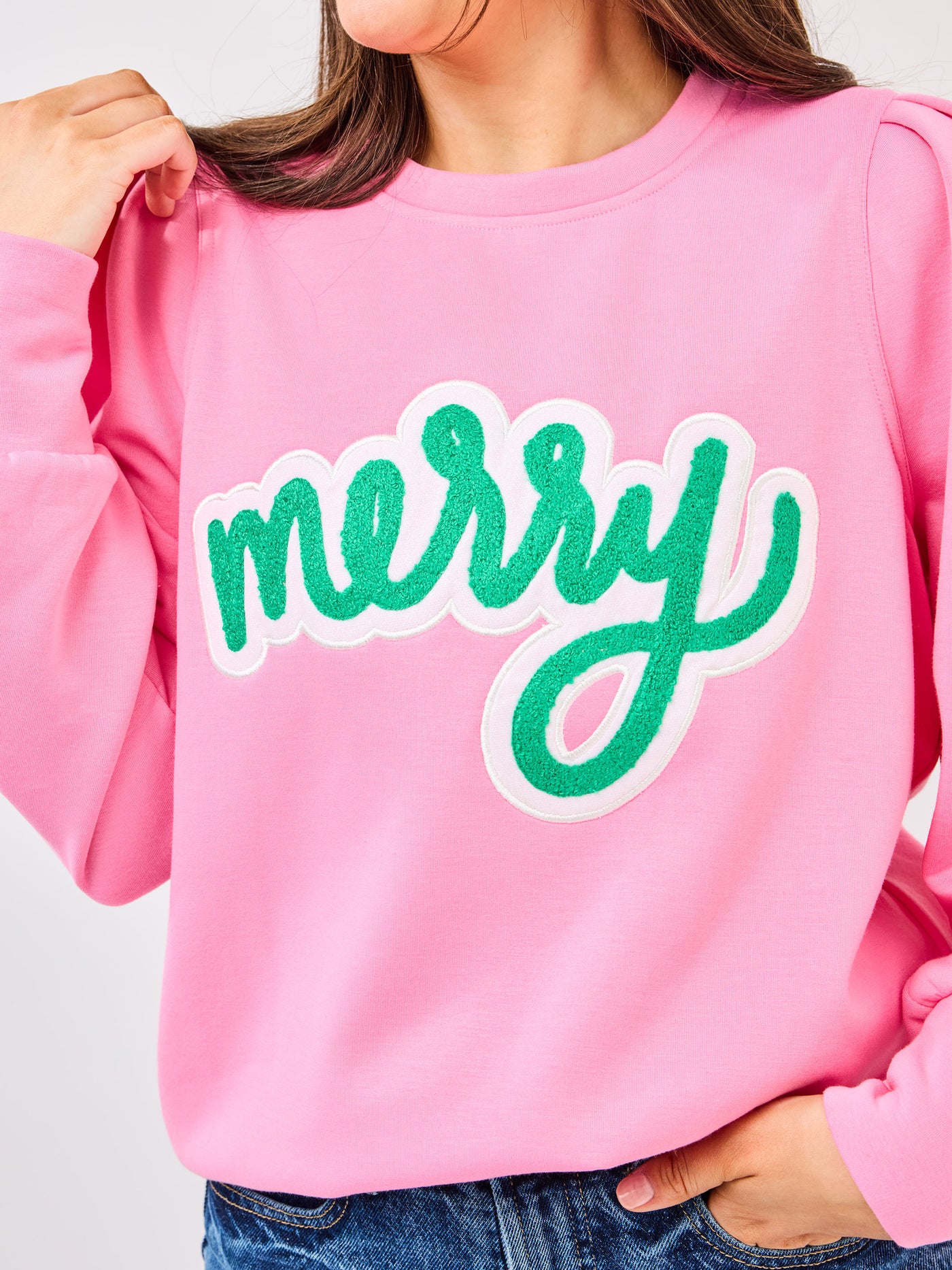 Person wearing a pink sweatshirt with 'merry' in green text on a plain background
