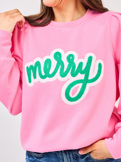 Person wearing a pink sweatshirt with 'merry' in green text on a plain background