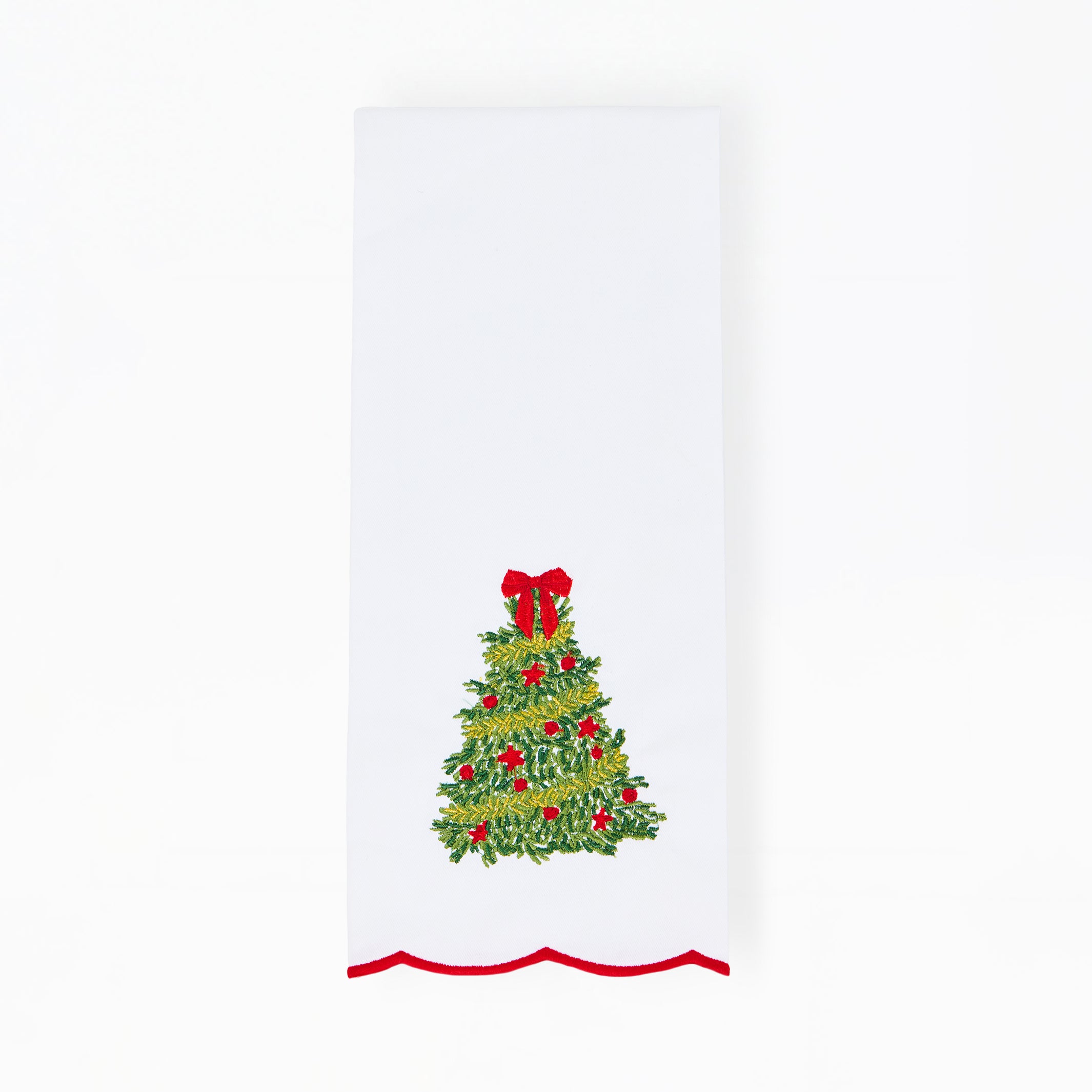 A white rectangular fabric item, likely a napkin or tea towel, featuring a green Christmas tree design with red ornaments and a red bow, finished with a red wavy scalloped trim along the bottom edge.