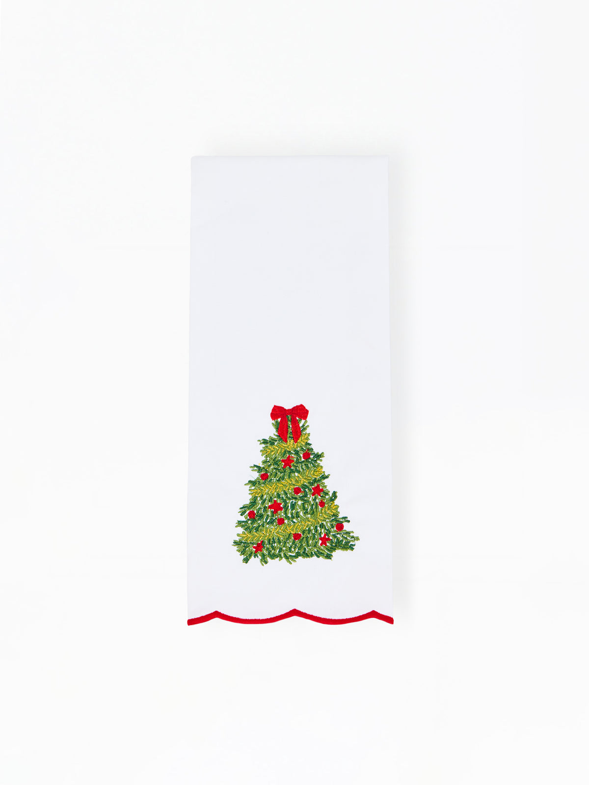 A white rectangular fabric item, likely a napkin or tea towel, featuring a green Christmas tree design with red ornaments and a red bow, finished with a red wavy scalloped trim along the bottom edge.