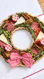 A holiday charcuterie food board arranged in a wreath shape on a scalloped wooden board, featuring sliced salami, cheese wedges, green olives, crackers, rosemary sprigs, and red-and-white gingham accents, with a large red-and-white striped bow at the bottom, photographed on a white surface.