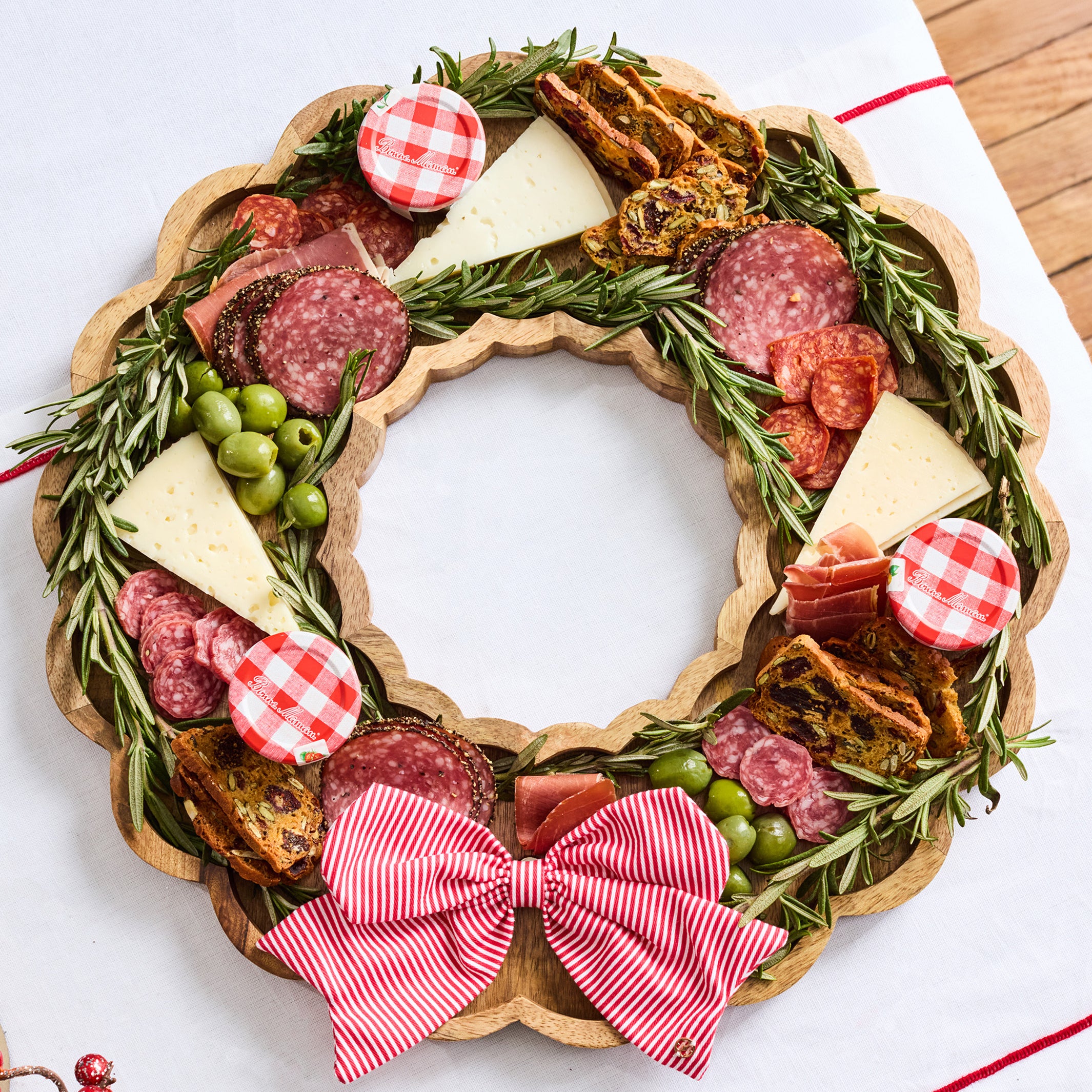 A holiday charcuterie food board arranged in a wreath shape on a scalloped wooden board, featuring sliced salami, cheese wedges, green olives, crackers, rosemary sprigs, and red-and-white gingham accents, with a large red-and-white striped bow at the bottom, photographed on a white surface.