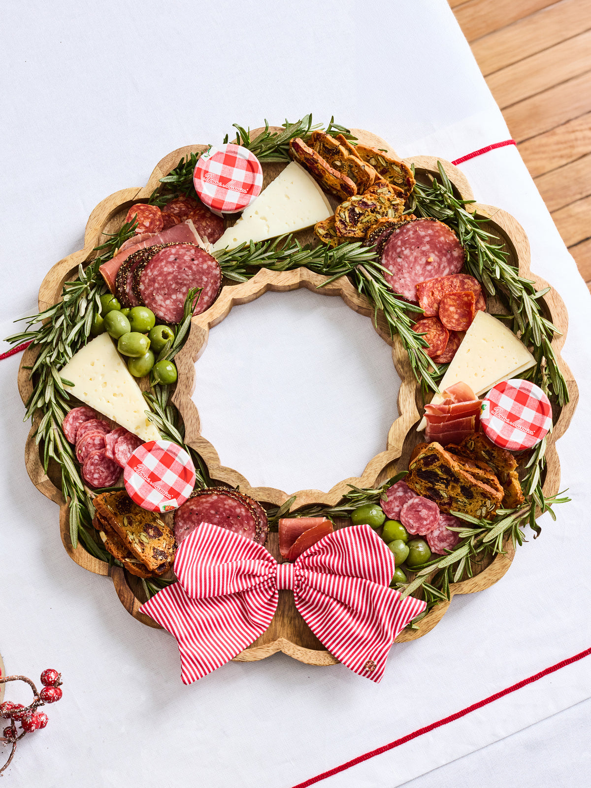 A holiday charcuterie food board arranged in a wreath shape on a scalloped wooden board, featuring sliced salami, cheese wedges, green olives, crackers, rosemary sprigs, and red-and-white gingham accents, with a large red-and-white striped bow at the bottom, photographed on a white surface.