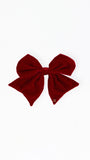 A large deep red velvet bow with a small gold logo charm on one tip, displayed flat against a solid white background.
