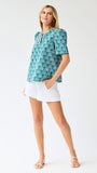 Woman wearing a teal patterned top and white shorts on a light gray background