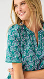 Woman wearing a teal patterned top and white shorts on a light gray background