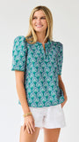 Woman wearing a teal patterned top and white shorts on a light gray background