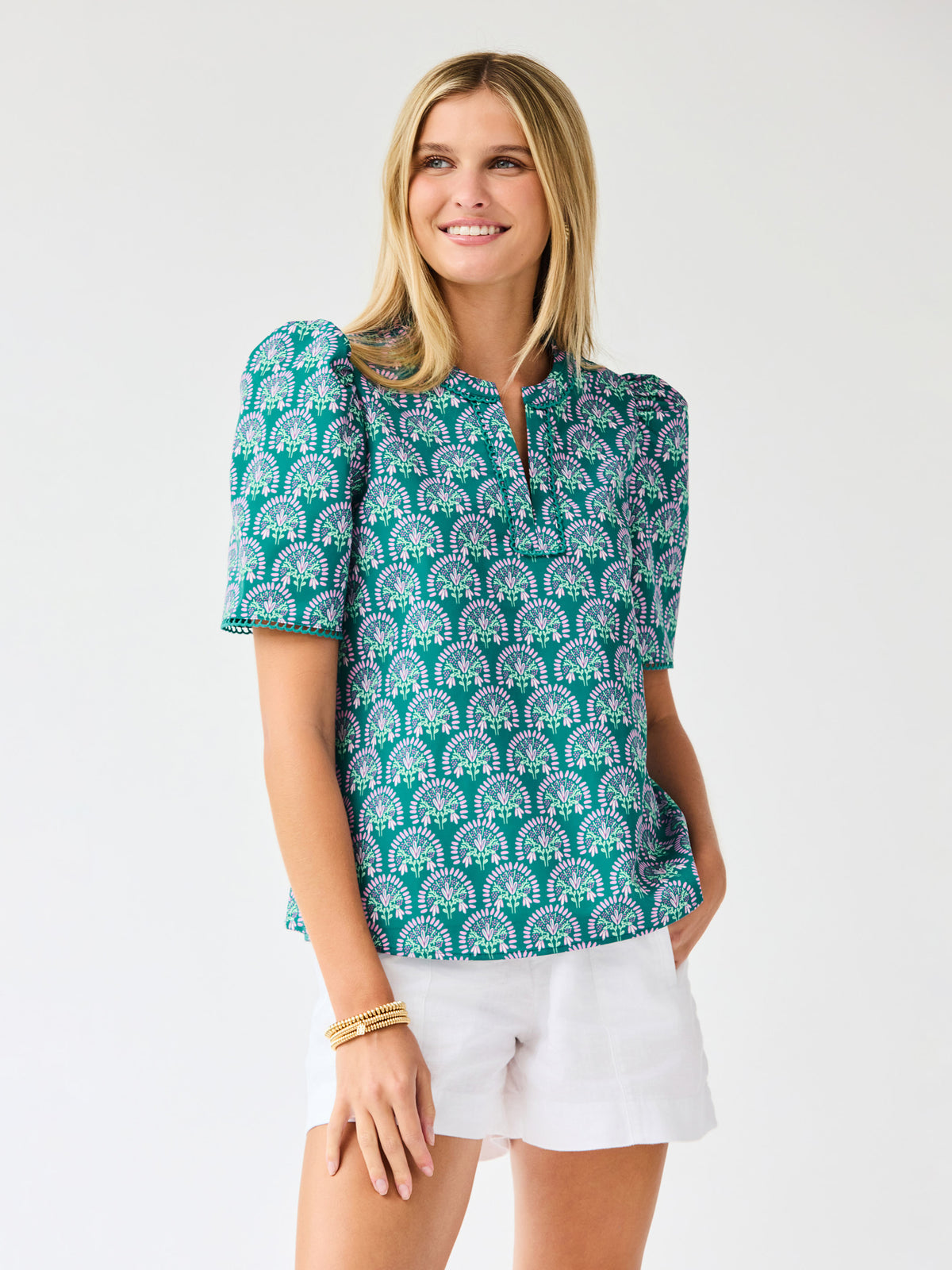Woman wearing a teal patterned top and white shorts on a light gray background