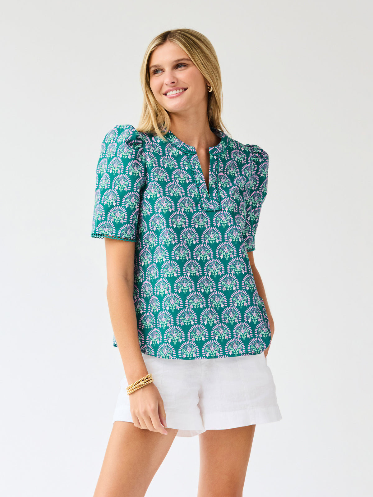 Woman wearing a teal patterned top and white shorts on a light gray background