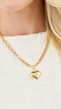 A close-up of a model wearing a gold chain necklace with a small, polished gold heart pendant, styled with a white textured top.