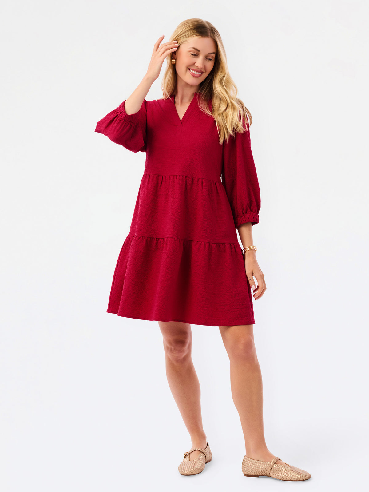 A woman modeling a tiered red dress with three-quarter puff sleeves and a V-neckline, paired with beige woven flat shoes, against a plain white studio background.