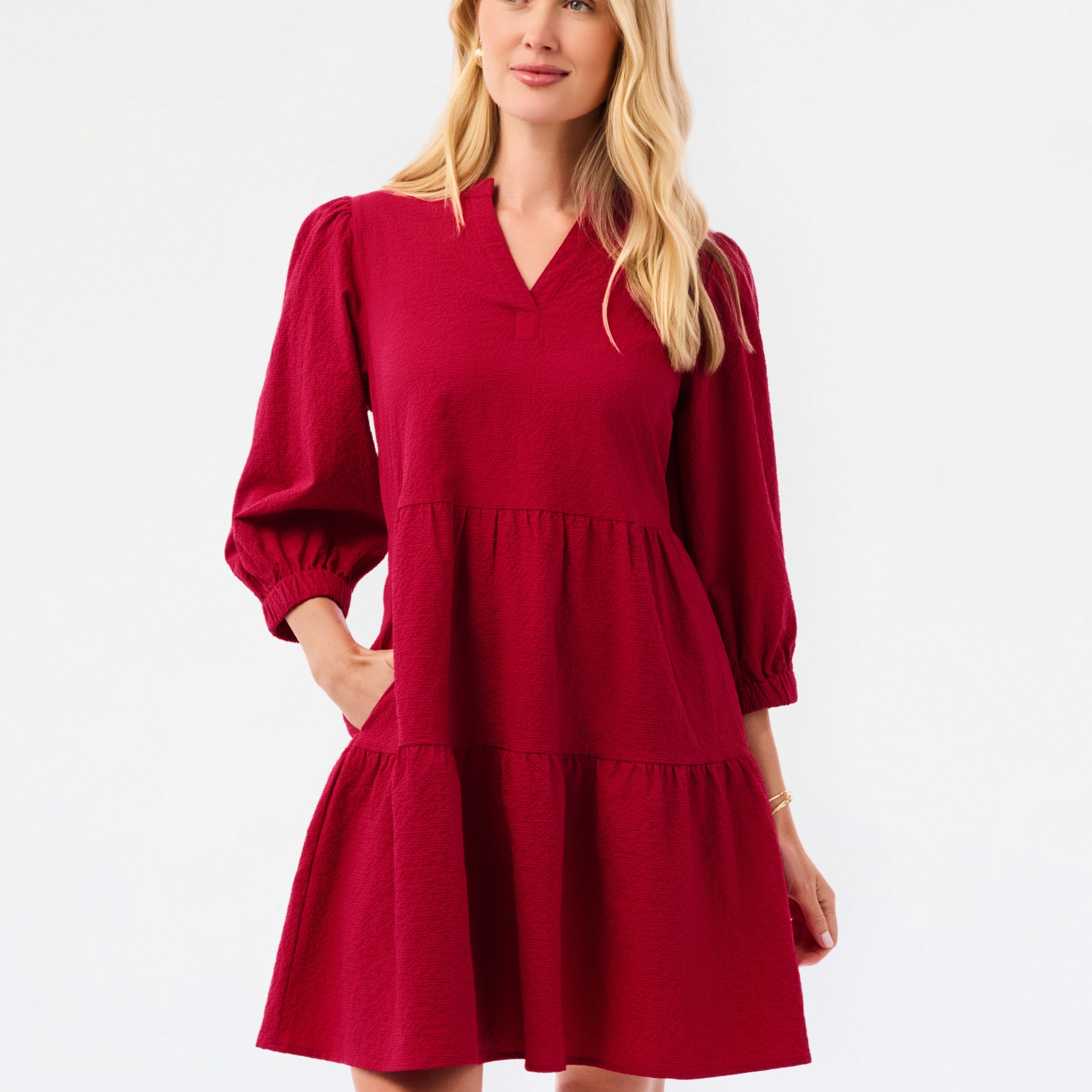 A woman modeling a tiered red dress with three-quarter puff sleeves and a V-neckline, photographed against a plain white studio background.
