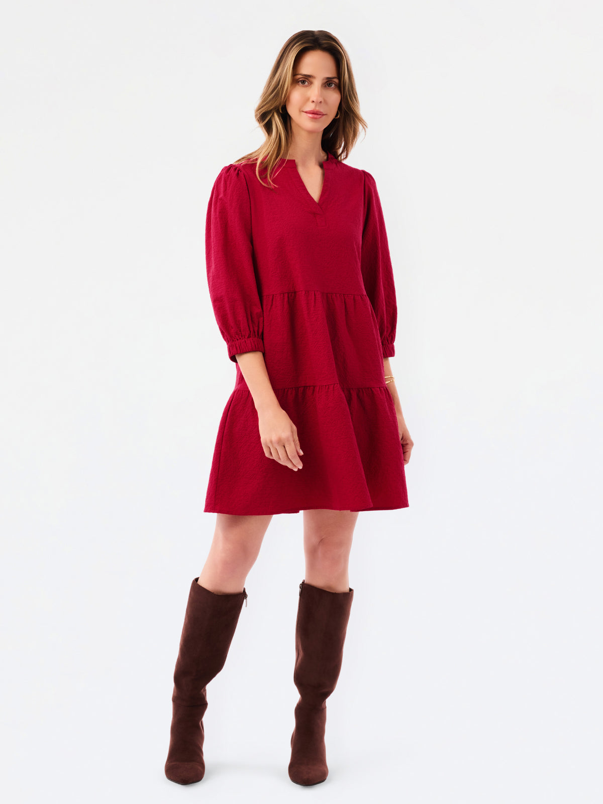 A woman modeling a short tiered red dress with three-quarter sleeves and brown knee-high boots, standing against a plain white studio background.