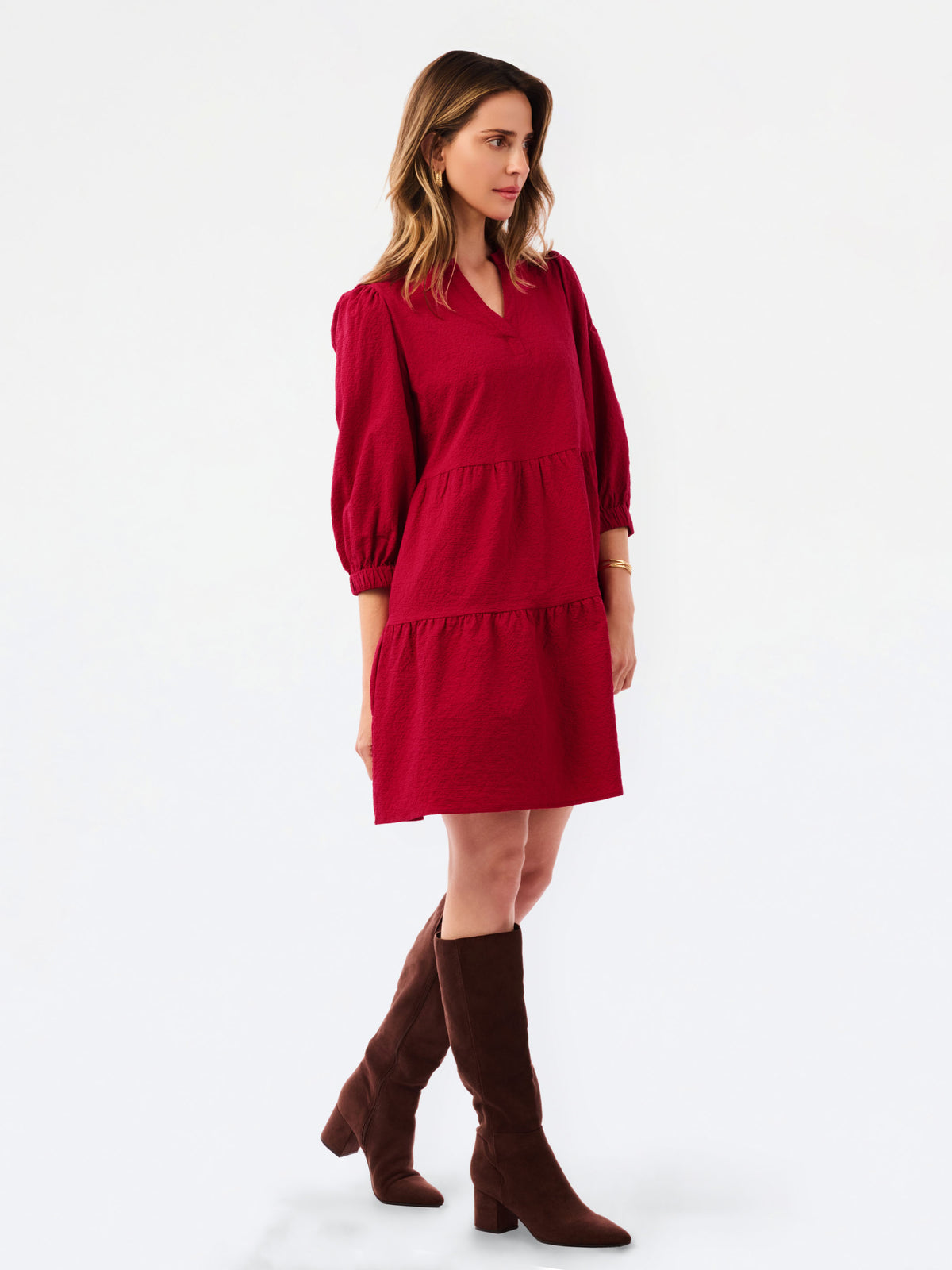 A woman modeling a red tiered mini dress with three-quarter puff sleeves, paired with dark brown knee-high boots, against a plain white studio background.