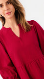 A woman wearing a deep red long-sleeve dress with a split V-neckline and gathered waist, shown in a close-up studio shot against a light gray background.
