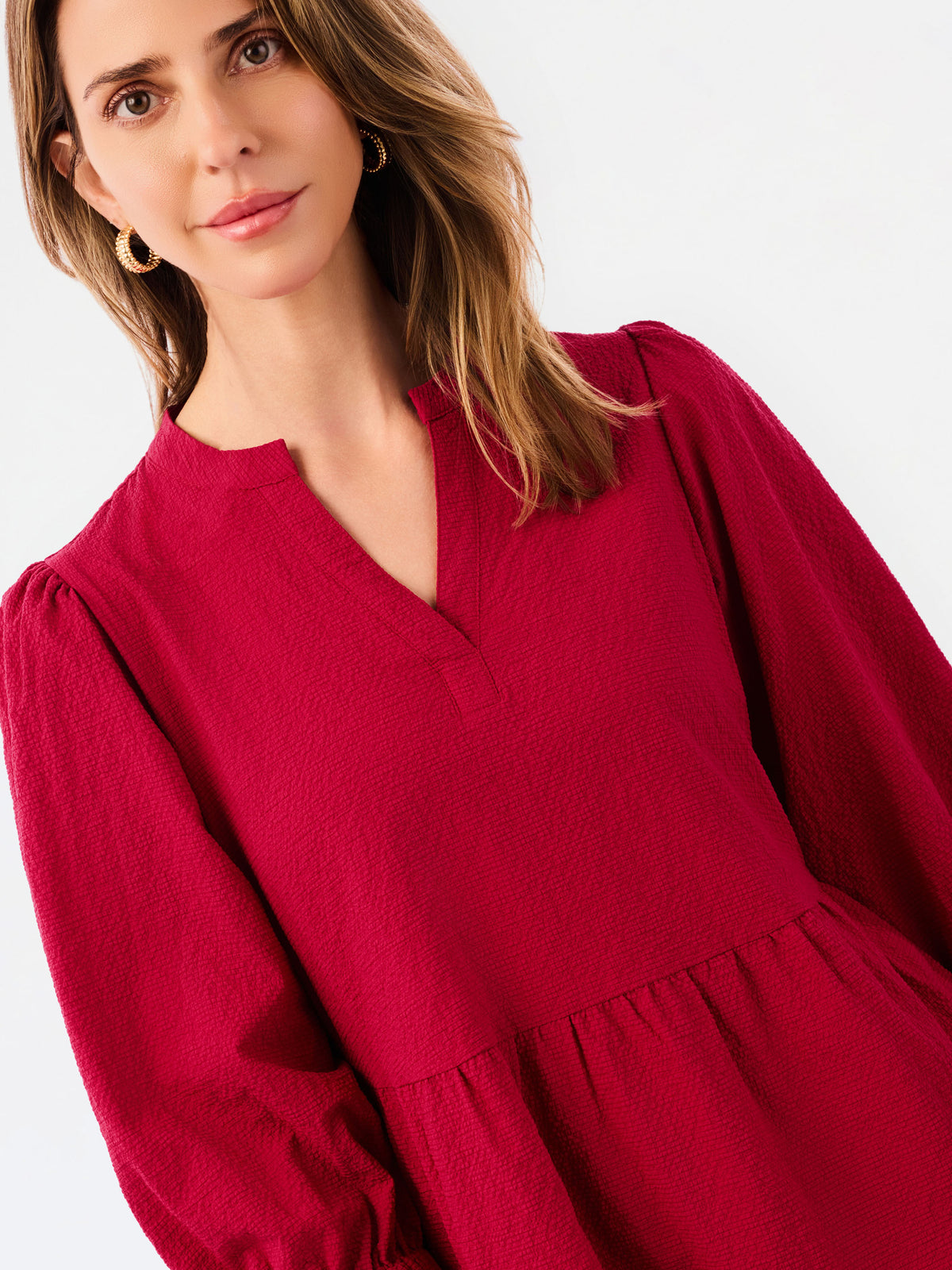 A woman wearing a deep red long-sleeve dress with a split V-neckline and gathered waist, shown in a close-up studio shot against a light gray background.