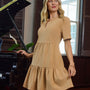A woman wearing a beige short-sleeve tiered dress stands indoors next to a black grand piano, with a window and dark wood shutters in the background.