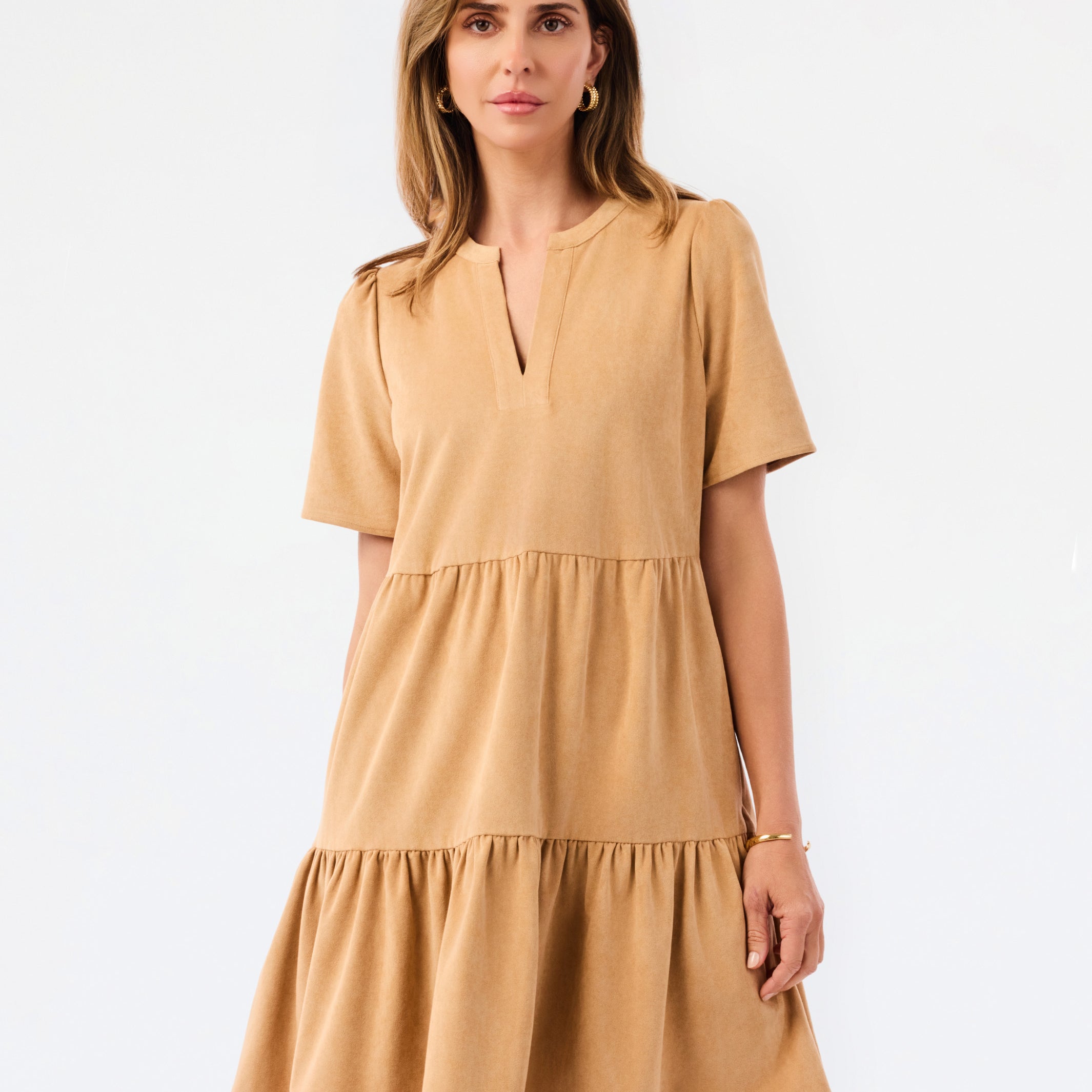 A woman modeling a short-sleeve tiered dress in a warm camel tan color, featuring a split neckline and ruffled hem, set against a plain white studio background.