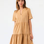 A woman modeling a short-sleeve tiered dress in a warm camel tan color, featuring a split neckline and ruffled hem, set against a plain white studio background.