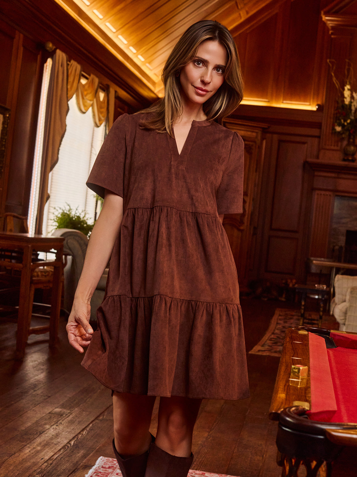 A woman wearing a brown tiered short-sleeve dress with a split neckline, standing in a warm, wood-paneled room.