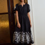 A woman modeling a black midi dress with white floral lace embroidery on the sleeves and hem, standing in an indoor setting next to a dark wooden door.