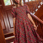 A woman wearing a short-sleeve, smocked-waist maxi dress with a red and pink floral print on a dark brown background, standing on a wooden staircase.