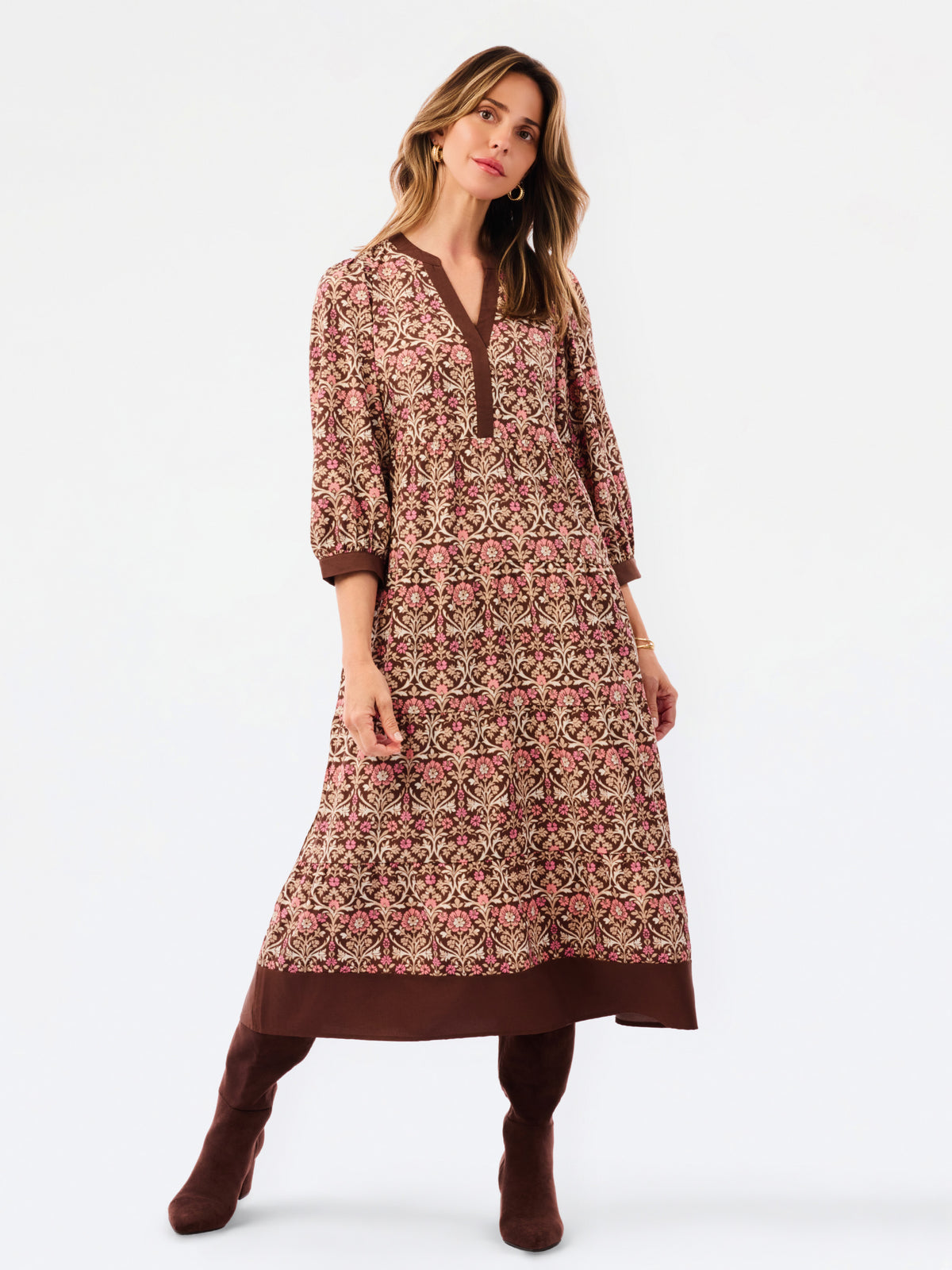 A woman modeling a long-sleeved, knee-length printed tunic dress in pink, brown, and beige, paired with matching brown boots, against a plain white studio background.
