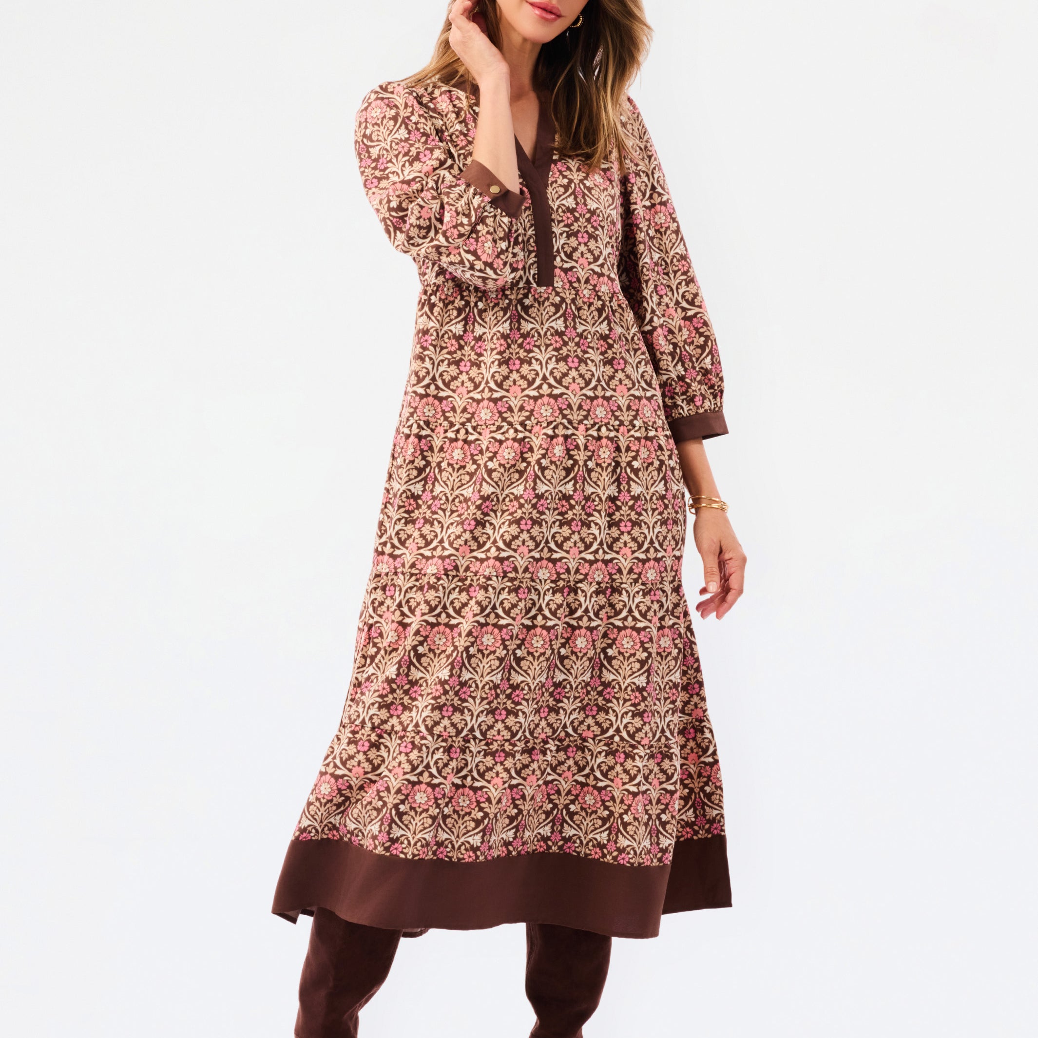 A woman modeling a long-sleeved, knee-length printed tunic dress in pink, brown, and beige, paired with matching brown boots, against a plain white studio background.
