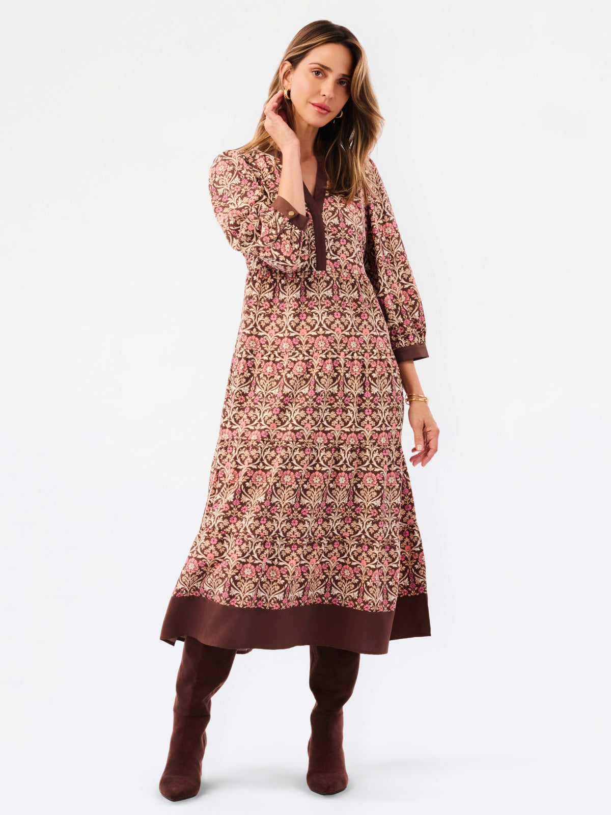 A woman modeling a long-sleeved, knee-length printed tunic dress in pink, brown, and beige, paired with matching brown boots, against a plain white studio background.